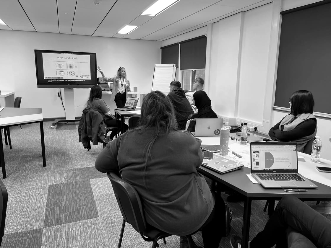 A presenter beside a screen reading 'What is inclusion?', speaking to a room of adult learners at tables with laptops and notes.