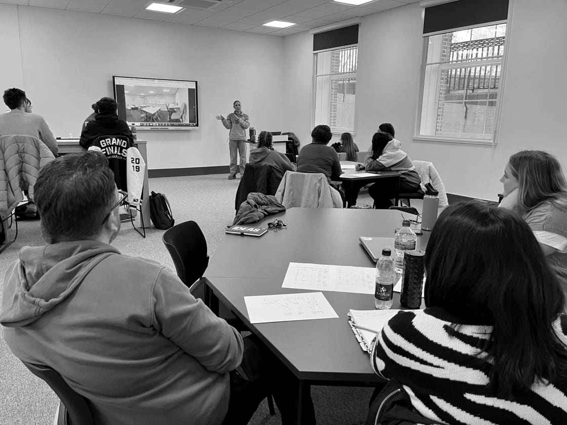 A ReFrame Inc session in progress: a presenter standing beside a wall screen, addressing adult learners seated around tables.