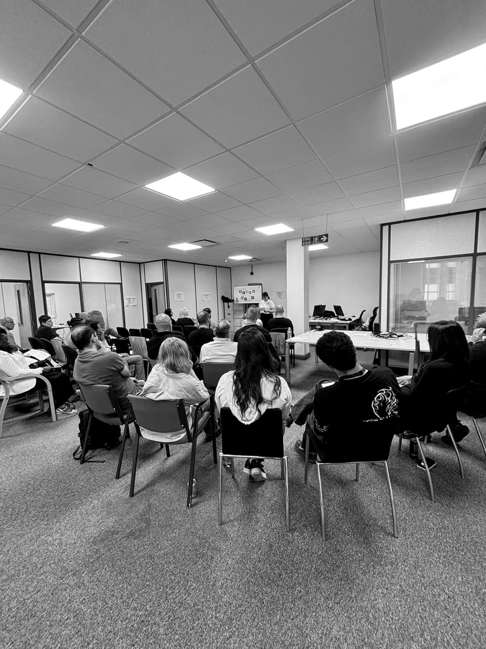 A training room seen from the back, every seat taken, adult learners facing a presenter at a wall screen.